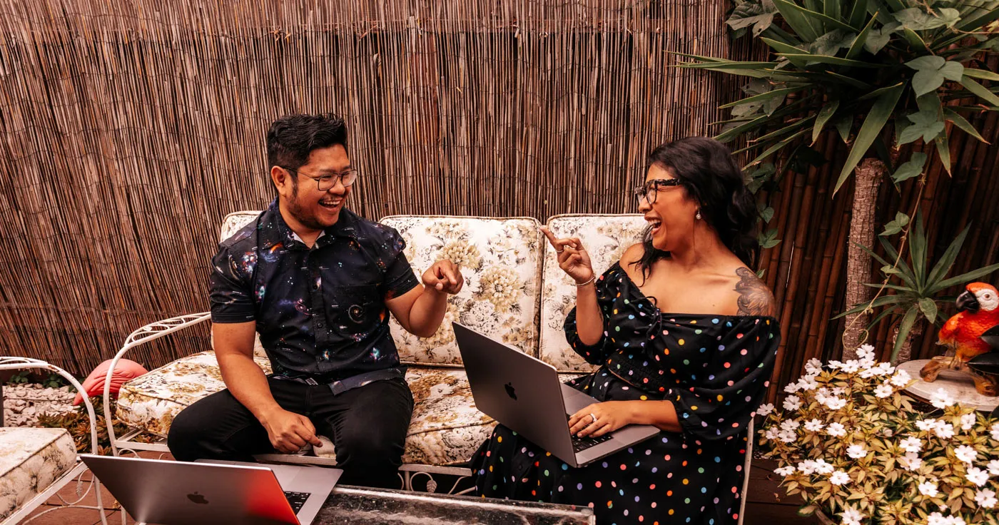 Jess and Nelson laughing outdoors with laptops on the floral couch